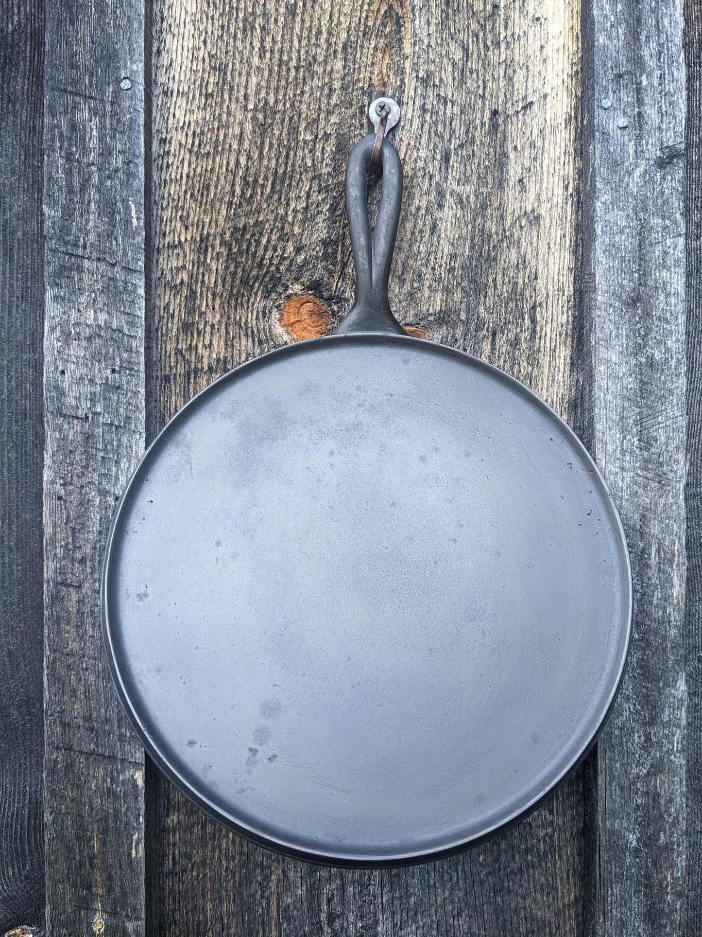 Cast iron round skillet griddle with handle, size #9, unknown maker. Probably dates from the 1800s. It is hanging from a small metal hook on a barn wood wall.