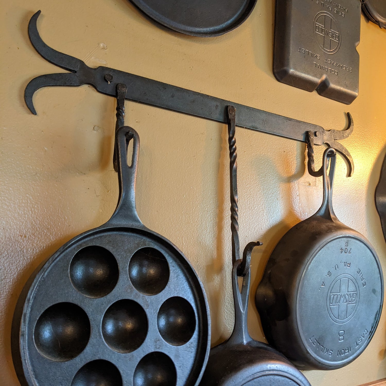 Custom three hook pot rack made by our blacksmith. Shown mounted on a tan wall holding vintage Griswold skillets.