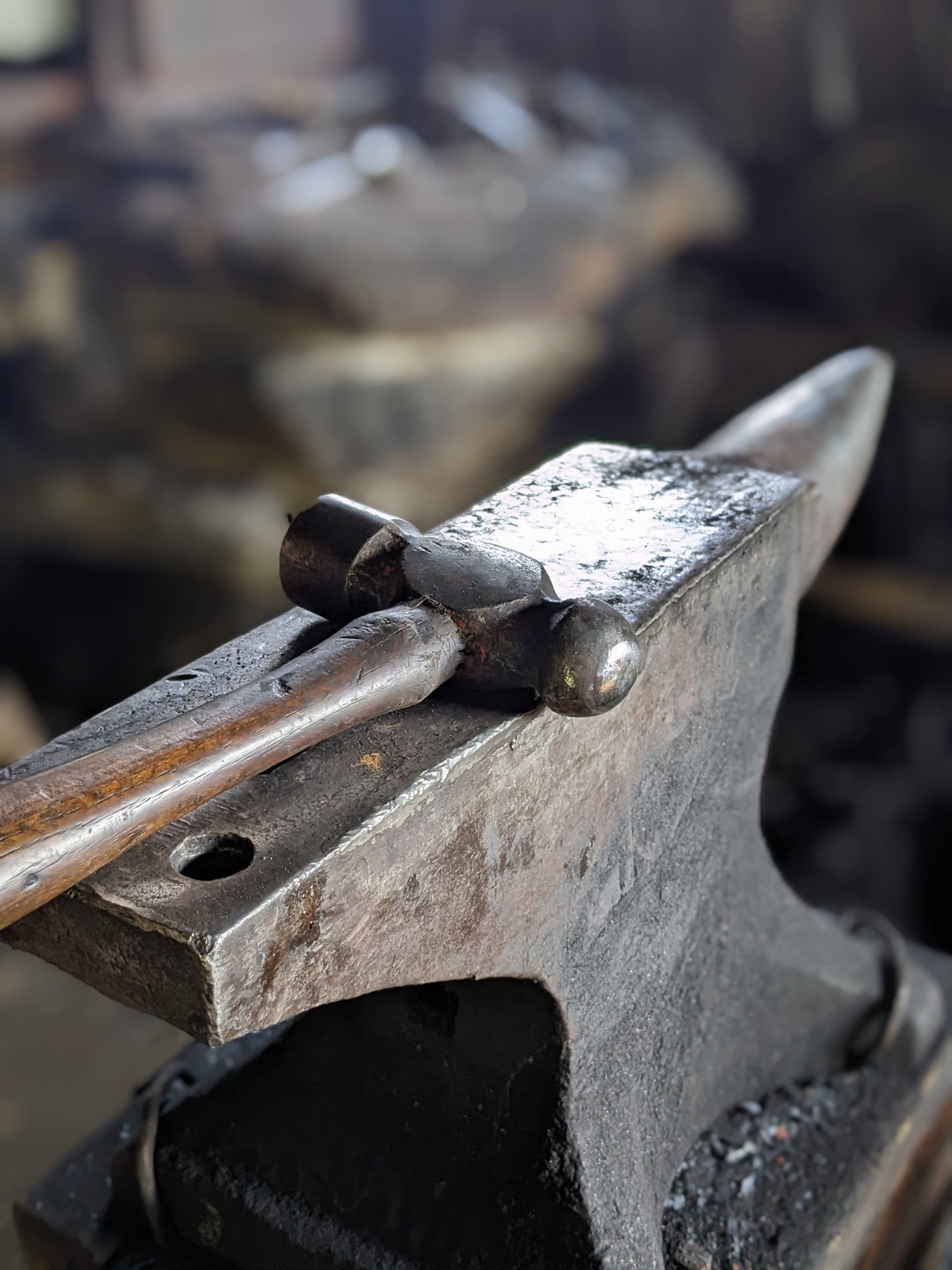A blacksmith's hammer sits on an anvil. The stone forge sits behind the anvil, slightly out of focus.  Our hand forged metal work is truly handmade.