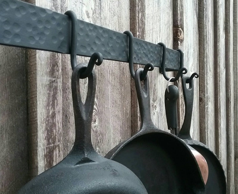 Side view of 48" hammer finish pot rack with the upgraded scroll end hooks shown. Hooks are shown holding cast iron pans and a copper bottom skillet. Mounted on a barn wood wall.
