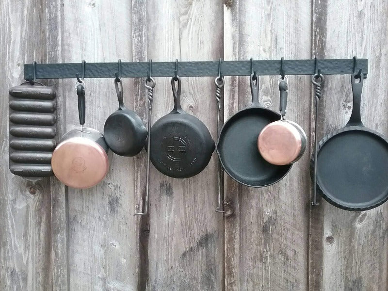 Front view of 48" hand forged hammer finish pot rack. Made by a blacksmith. Shown mounted onto a barn wood wall with cast iron pans, copper bottom pots and hand forged steak turners.