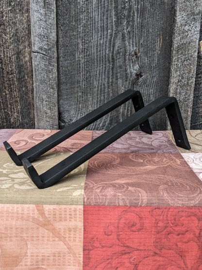 Heavy duty steel floating shelf brackets made by a blacksmith. they hold a 10" shelf and are shown on a red, grey and brown tablecloth in front of a barn wood wall.