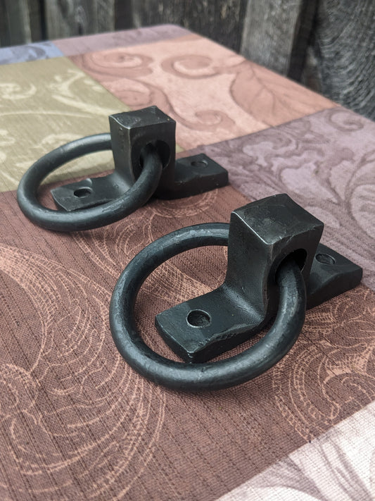 Two small hand forged ring handles sitting on a colorful tablecloth in front of a barn wood backdrop.