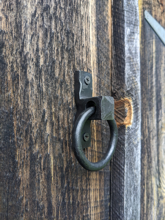 A single blacksmith made ring handle, measuring about 2.25" in diameter, is mounted to barn wood.
