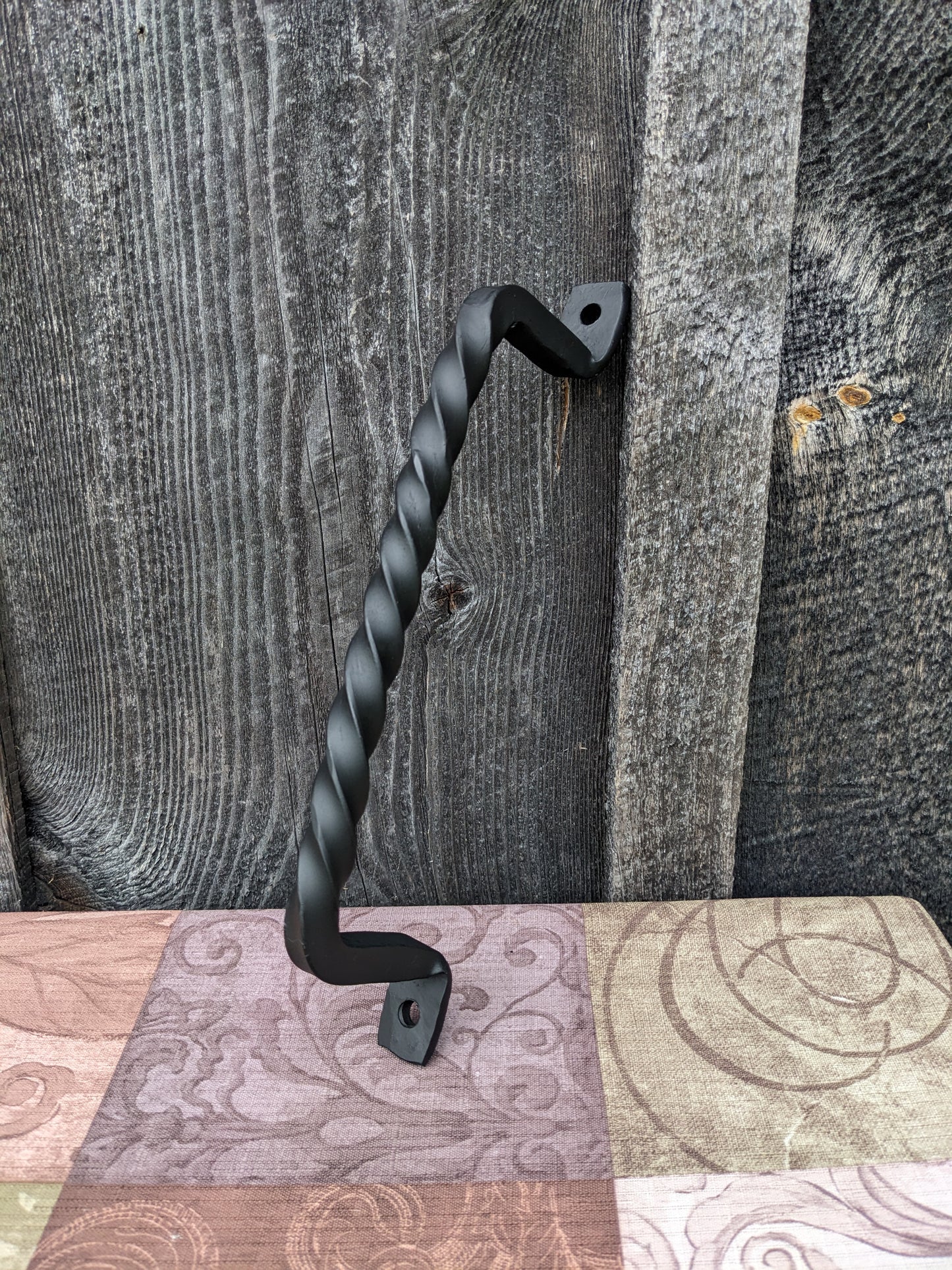 Hand Forged twisted handle with one end sitting on a table with a colorful tablecloth, the other side leaning against a barn wood wall.