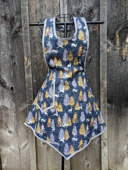 Apron made from a vintage 1940's pattern . Made from dark blue fabric with yellow and gray pine trees, white snowflakes and silver reindeer silhouettes printed all over. Fabric looks like a dreamy winter forest at twilight! Apron is shown on a wire frame showing off the feminine fit and pointed front hem.