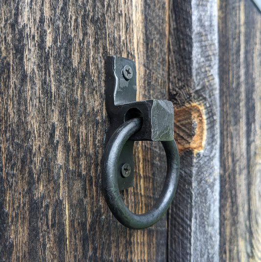 A single blacksmith made ring handle, measuring about 2.25" in diameter, is mounted to barn wood.