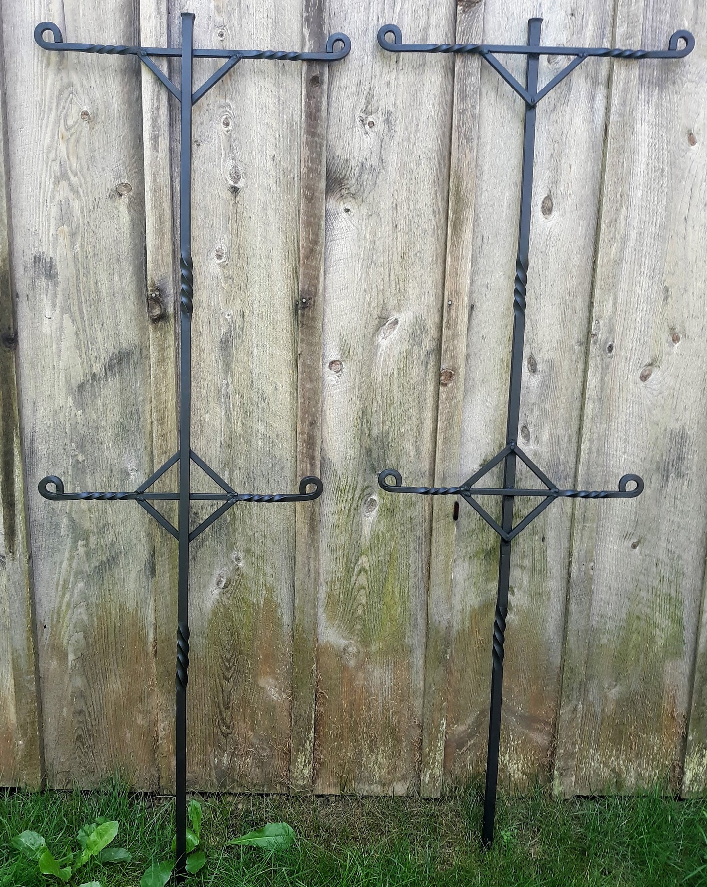 Two black hand forged trellis supports against a barnwood background.  Eah will hold up to 4 strands of trellis wire or rope.